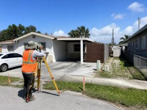 Surveyor in an orange safety vest uses a tripod mounted instrument in a residential driveway in front of a single-story house.