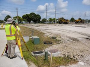 Construction worker in a neon safety vest uses a tripod surveying instrument at an active construction site with dirt and distant heavy machinery.